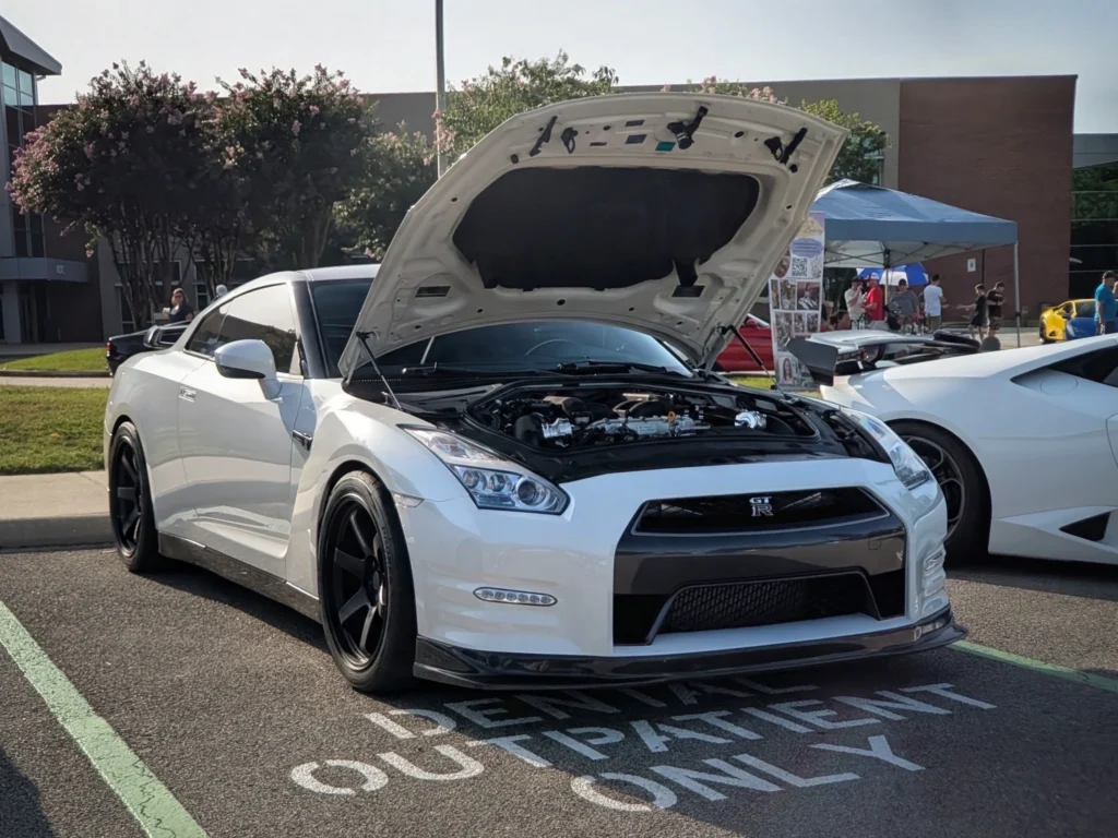 White Nissan GT-R with hood open showing engine at outdoor car event with tents and people in background