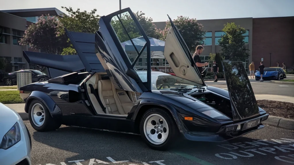 Black Lamborghini Countach 5000 QV with scissor doors open at a car show, beige interior visible, parked outdoors with other cars and