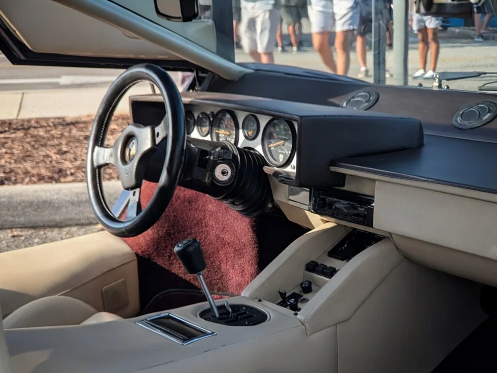 Beige Lamborghini Countach 5000 QV interior with black steering wheel and dashboard gauges visible, people walking outside through open door