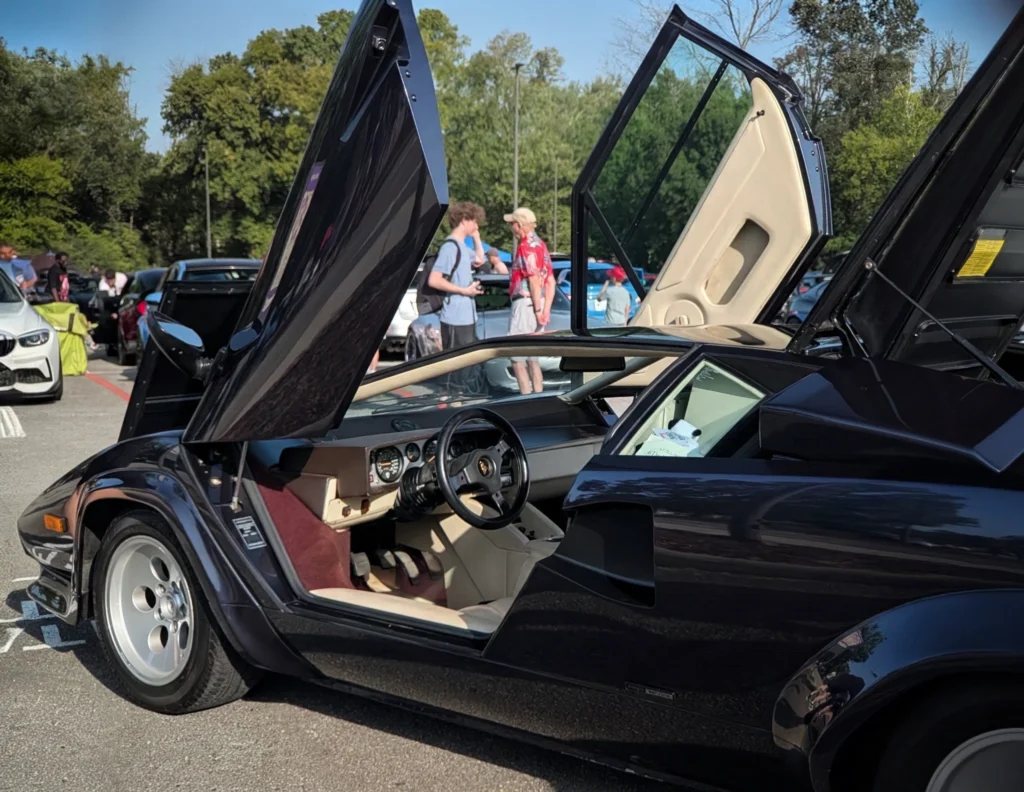Black Lamborghini Countach 5000 QV with scissor doors open showing interior at outdoor car event with people and trees in background