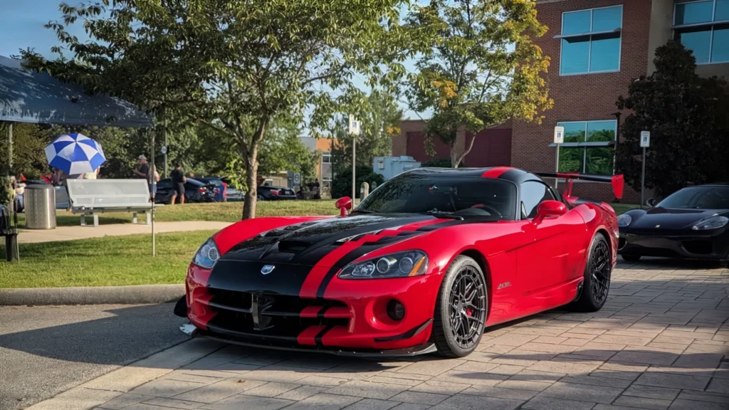 Red Dodge Viper ACR with black racing stripes parked on pavement near a building and trees at a car event