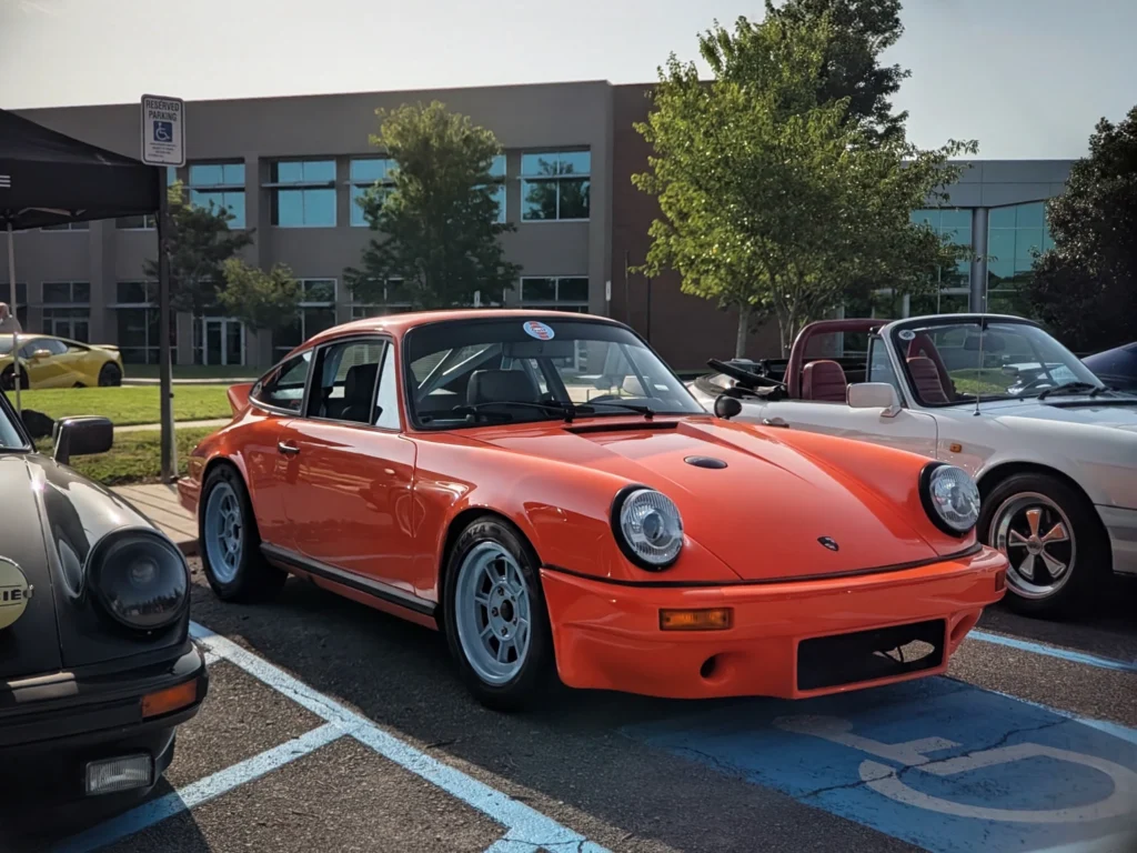 Orange Porsche 911 parked in a lot next to a white convertible and other cars at an outdoor car event.
