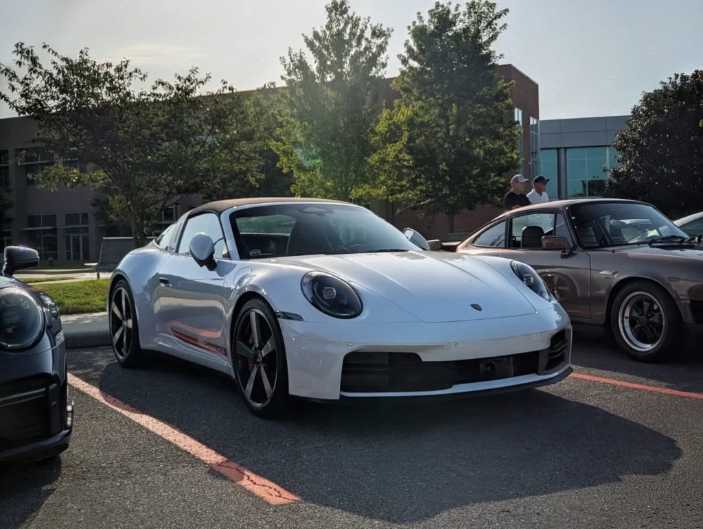 White Porsche 911 Targa 4S parked outside near trees and a building with people standing beside a dark-colored coupe