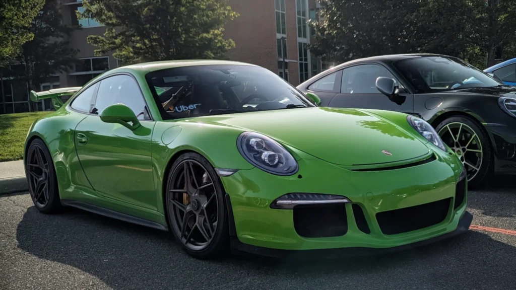Green Porsche 911 GT3 parked alongside black Porsche 911 in an outdoor parking lot with trees and buildings in background