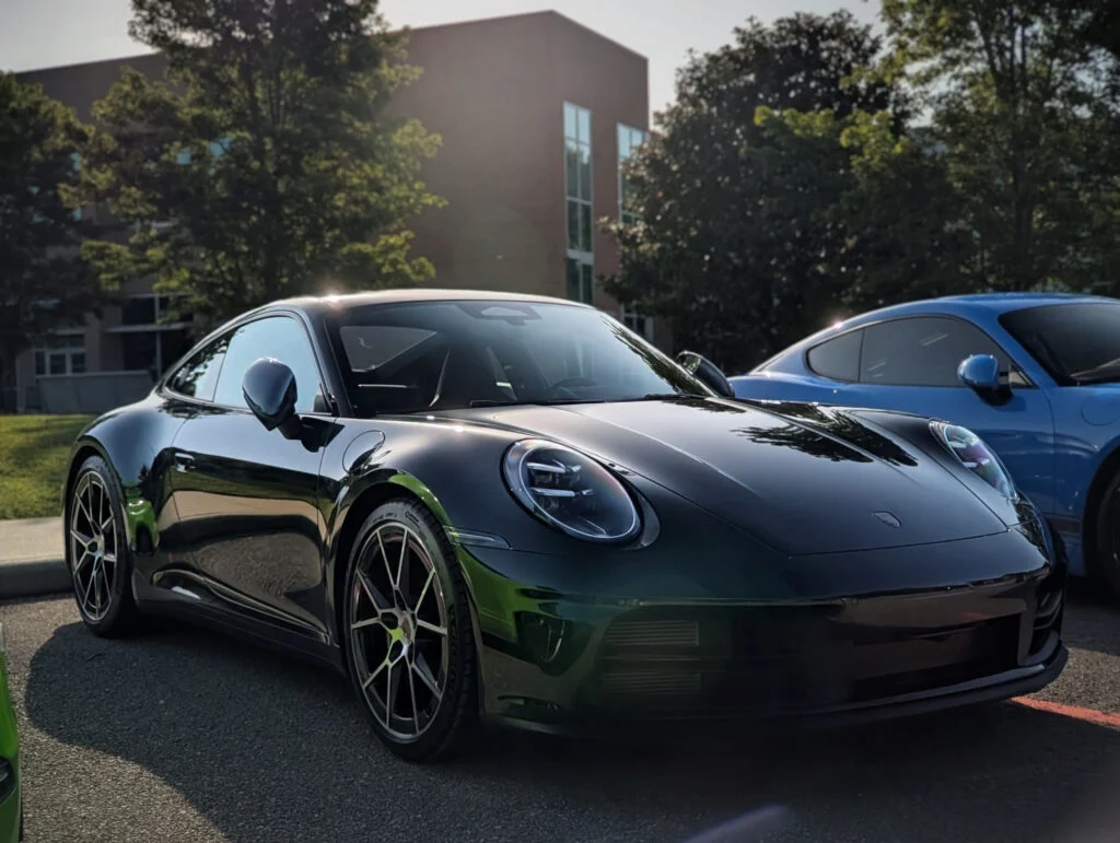 Black Porsche 911 parked outdoors near another blue Porsche with trees and building in the background