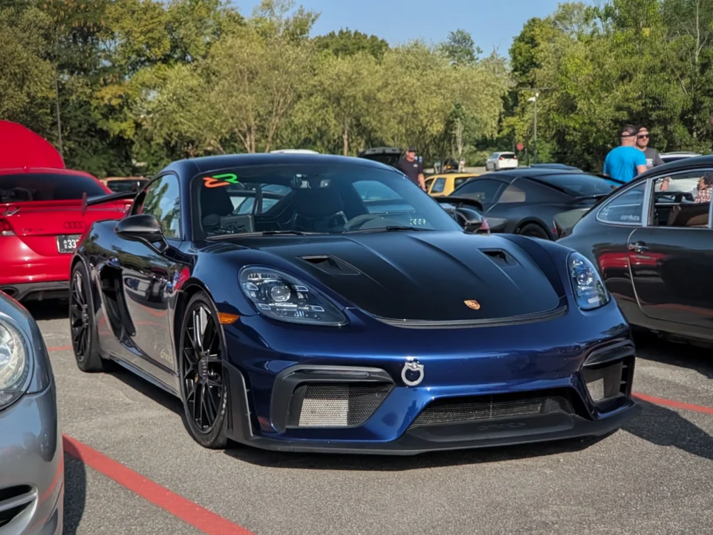 Dark blue Porsche 718 Cayman GT4 RS front 3/4 with other cars and people in parking lot at car event
