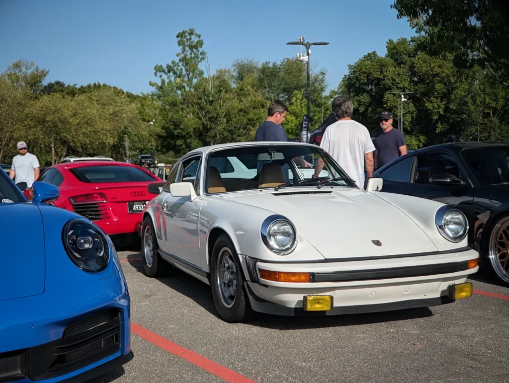 White Porsche 911 parked among other cars at an outdoor event with people standing nearby under clear blue sky.