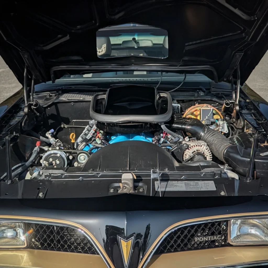 Engine bay view of a black and gold Pontiac Trans Am with hood open, revealing components and part of the interior through a rear wind