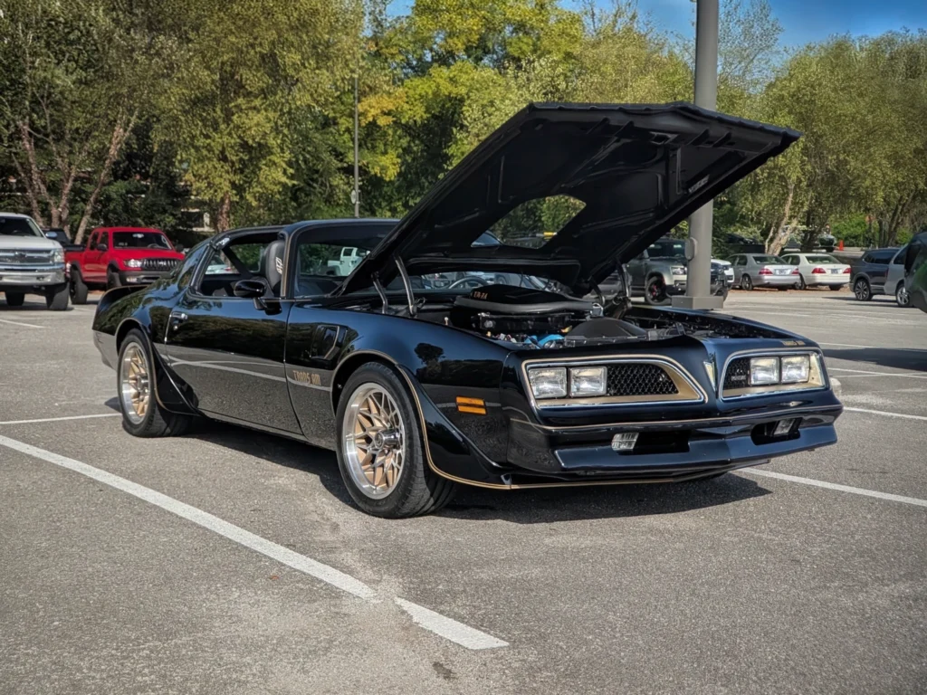 Black Pontiac Trans Am with hood open in a parking lot at a car event, surrounded by other vehicles and trees in background.