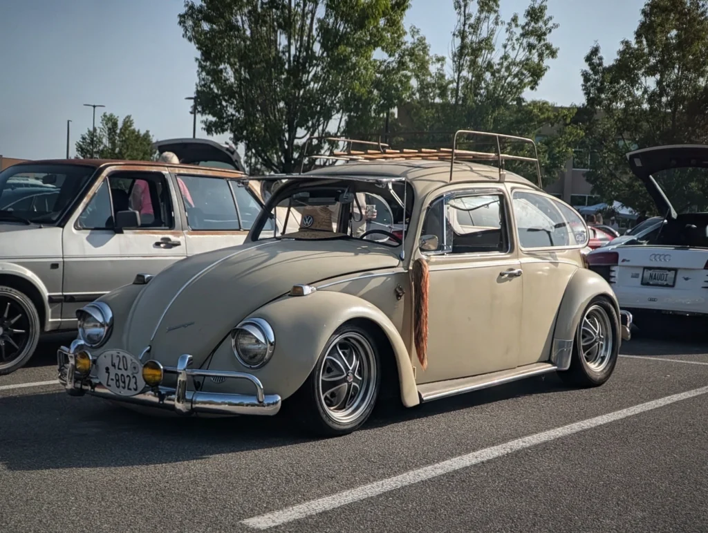 Light beige Volkswagen Beetle with roof rack and open side window in parking lot with other cars and trees.