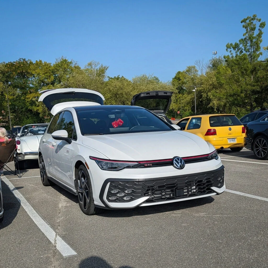 White Volkswagen GTI with open rear hatch parked in a lot with other cars and trees in the background