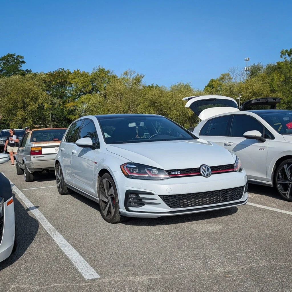White Volkswagen Golf GTI parked in a lot with other cars and green trees in the background under a clear blue sky.