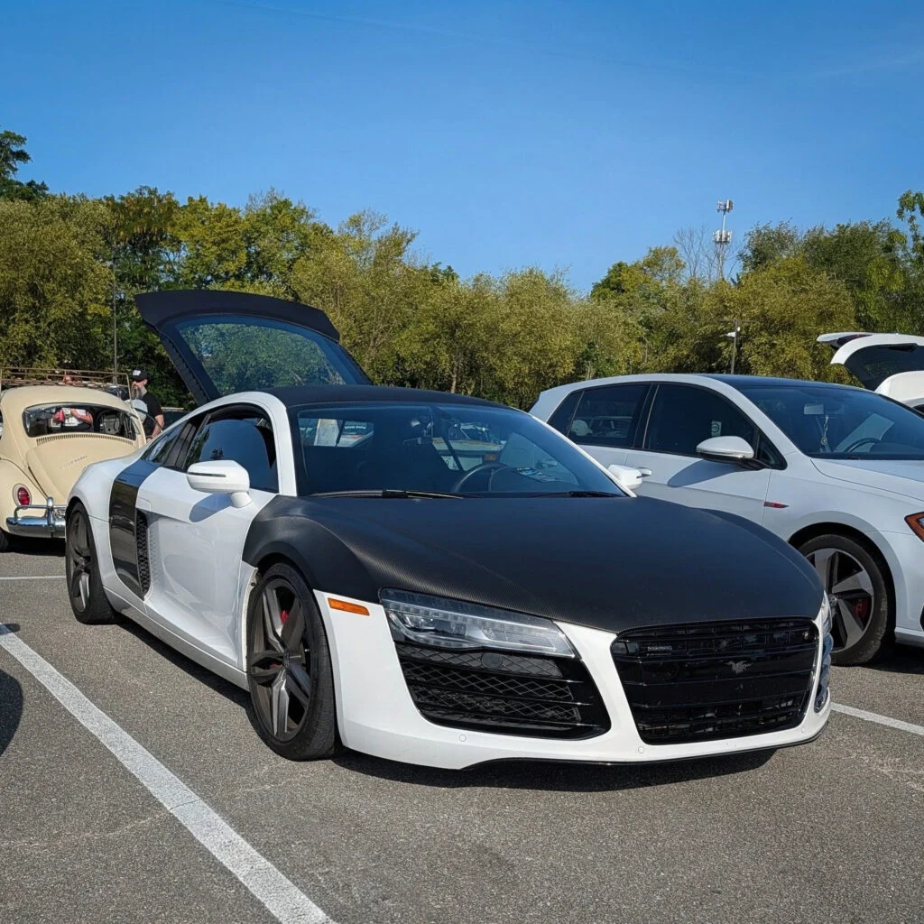 White Audi R8 with a black hood parked with its driver's side door open in an outdoor car event lot on a clear day