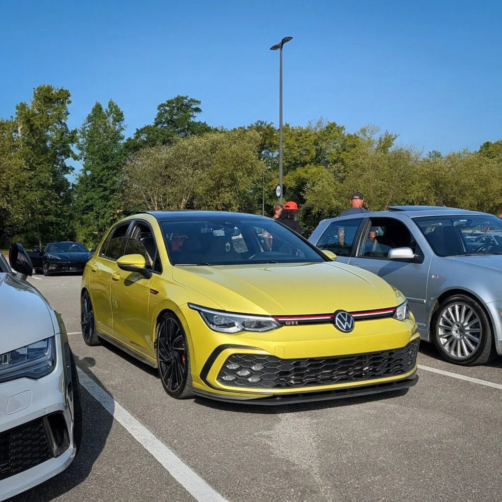 Yellow Volkswagen GTI parked alongside other cars in an outdoor parking lot with people and trees in the background.