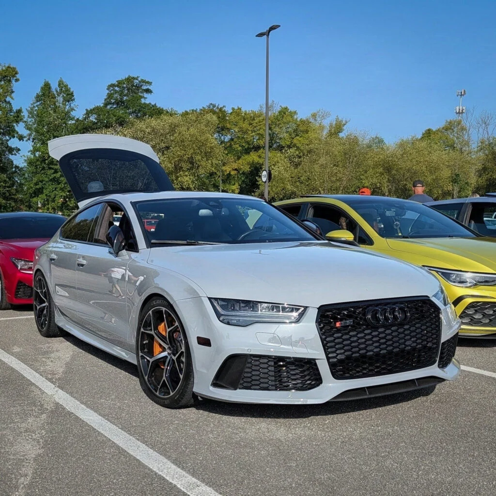 Silver Audi RS7 with rear hatch open parked next to a yellow SUV in an outdoor car event parking lot under clear sky