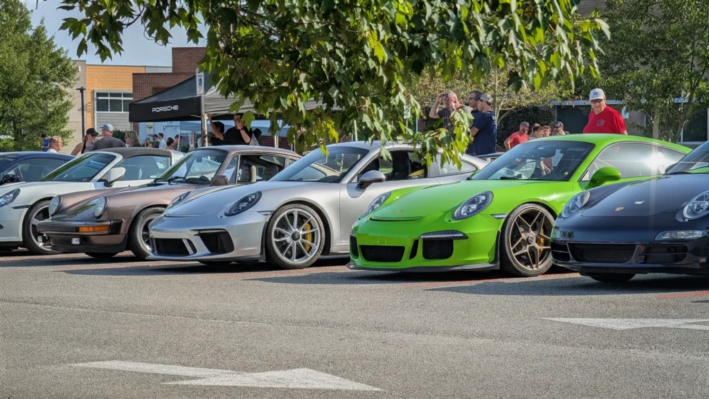 Several Porsche sports cars parked side by side at an outdoor car event with people in the background