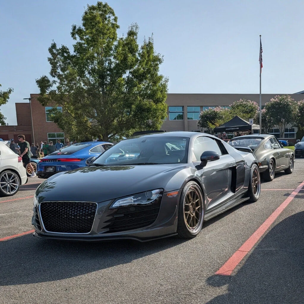 Black Audi R8 parked beside a blue Porsche 911 and a silver classic car at a daytime car event with people and buildings in t