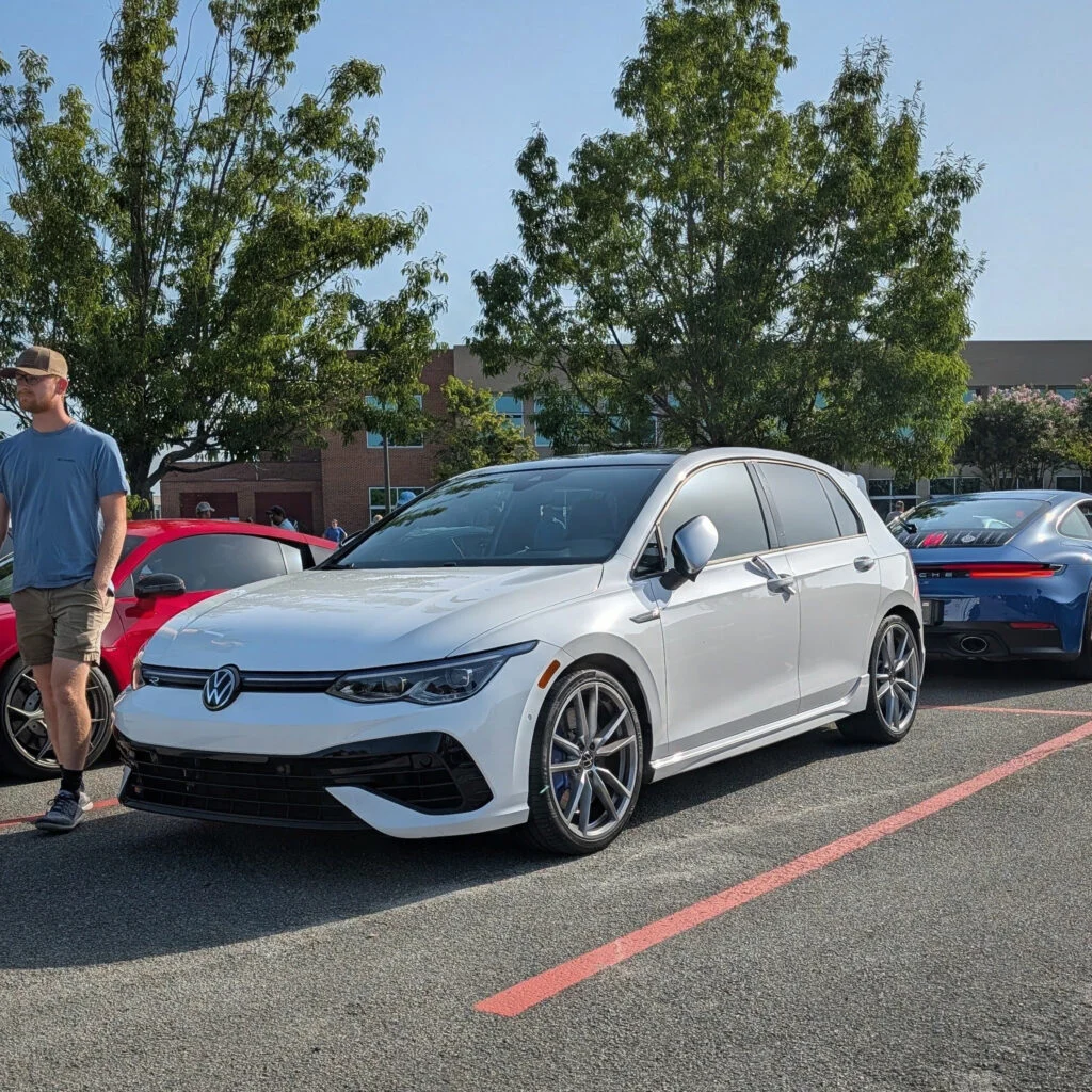 White Volkswagen hatchback parked at an outdoor car event with other vehicles and people in the background.