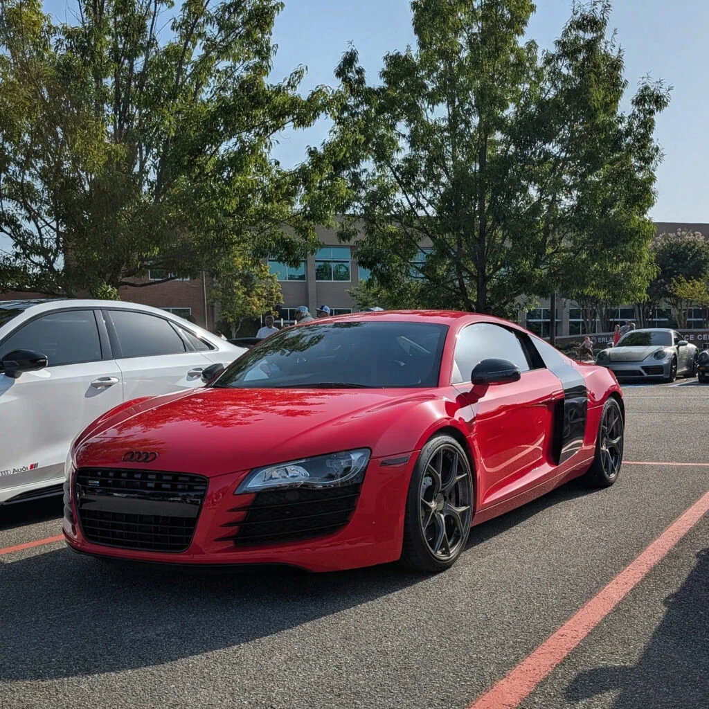 Red Audi R8 and white Audi sedan parked side by side in a parking lot at a daytime car event with people and trees in backgro