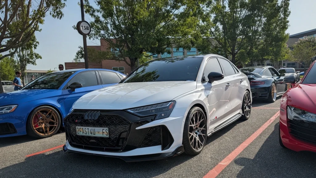 White Audi RS3 parked alongside a blue Audi and black Porsche in an outdoor car event parking lot with trees and people in ba