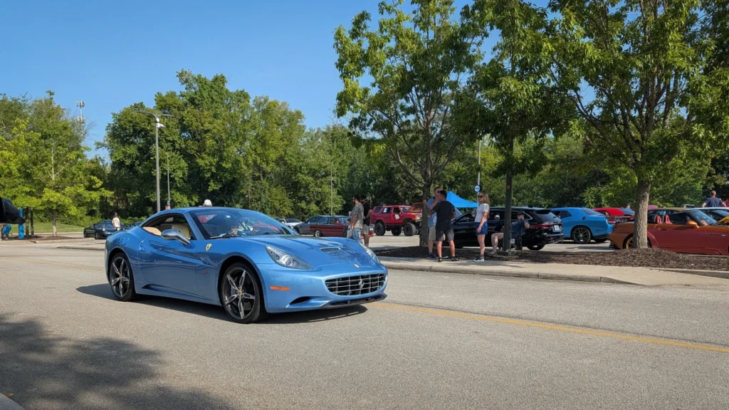 Light blue Ferrari California parked on road with trees and people in background at car event