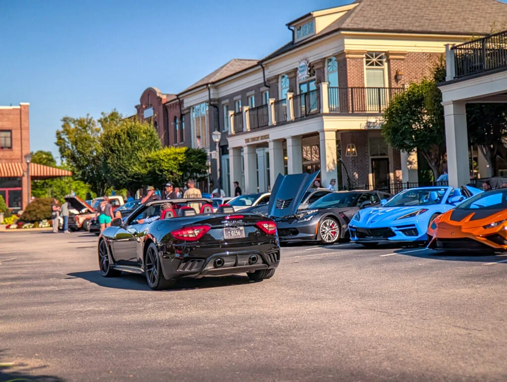Black Maserati convertible driving past a row of parked sports cars and people near retail buildings on a clear day