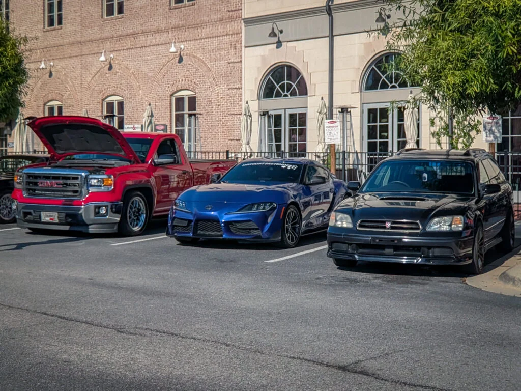 Red GMC pickup with open hood, blue Toyota Supra, and black Subaru Legacy parked in front of brick and stone buildings
