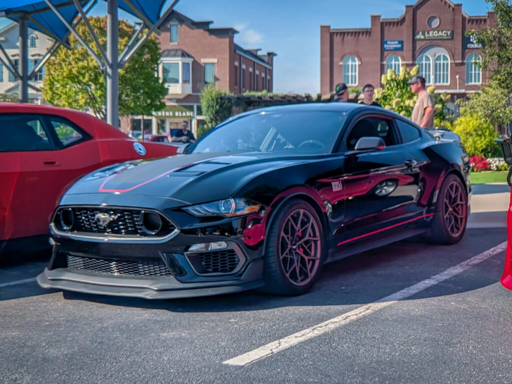 Black Ford Mustang Mach-1 parked beside a red car in an outdoor car event with people and buildings in the background