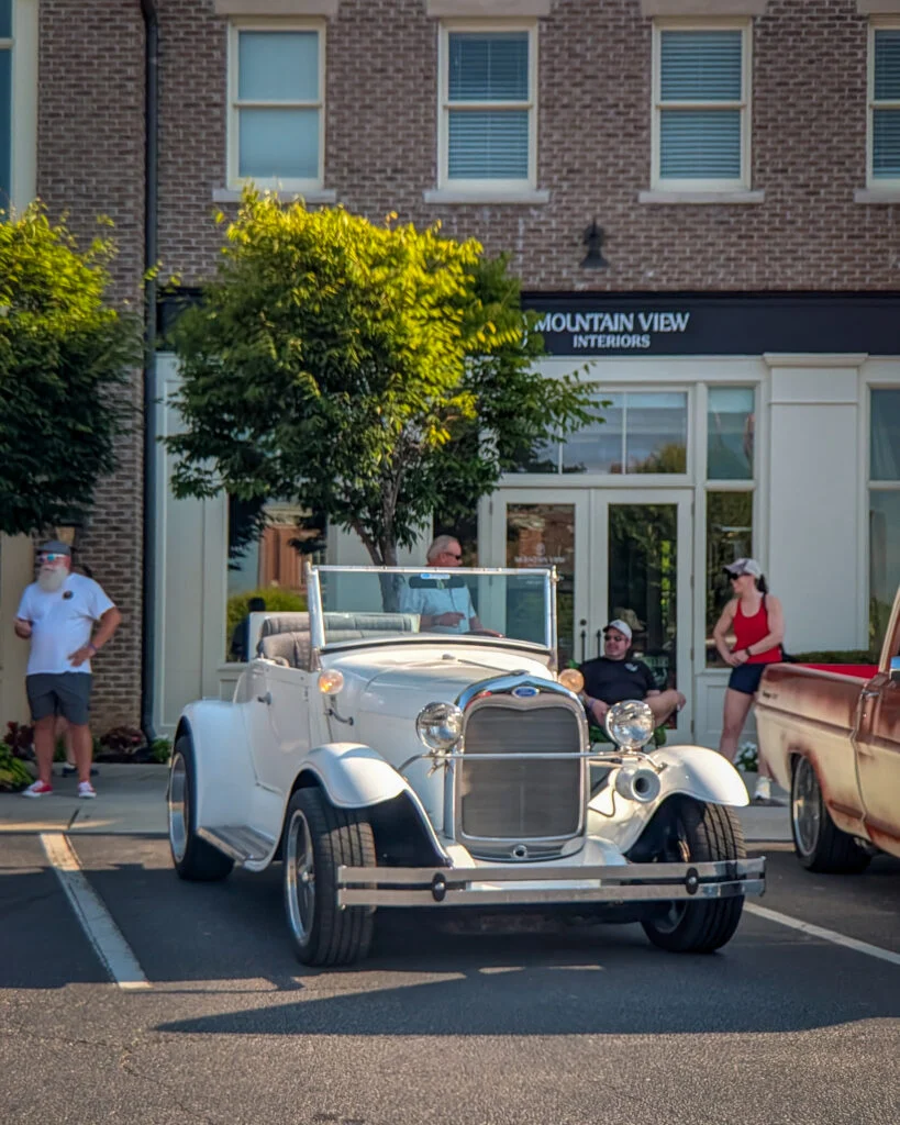 White vintage Ford Model A Roadster parked in front of a building with people standing nearby