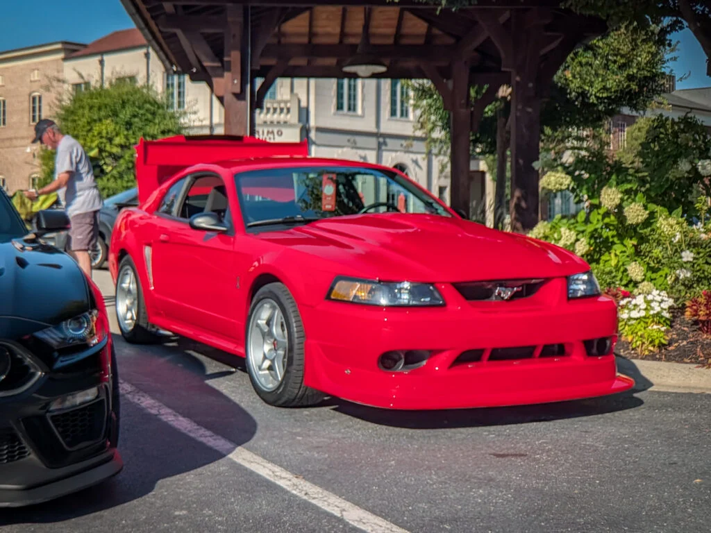 Red Ford Mustang SVT Cobra R with hood raised parked in a lot near a wooden shelter and greenery on a sunny day
