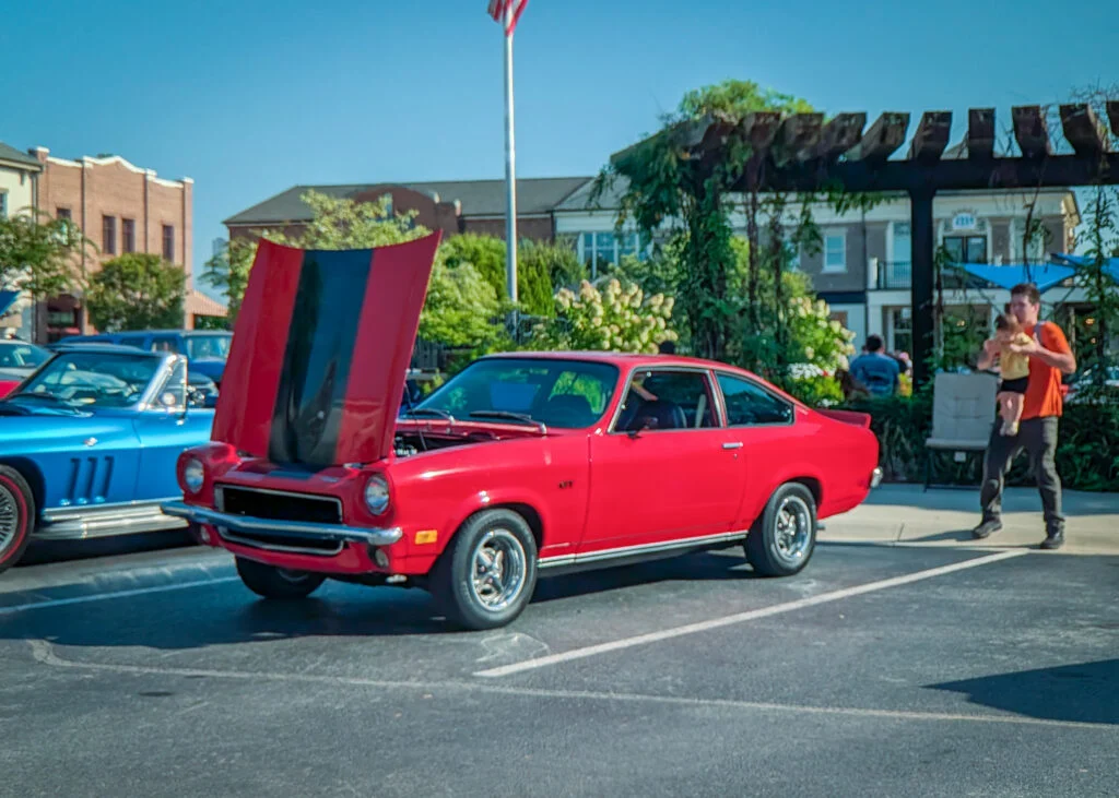 Red Chevrolet Vega with raised hood showing black stripe, parked beside a blue convertible at a car meet on a sunny day