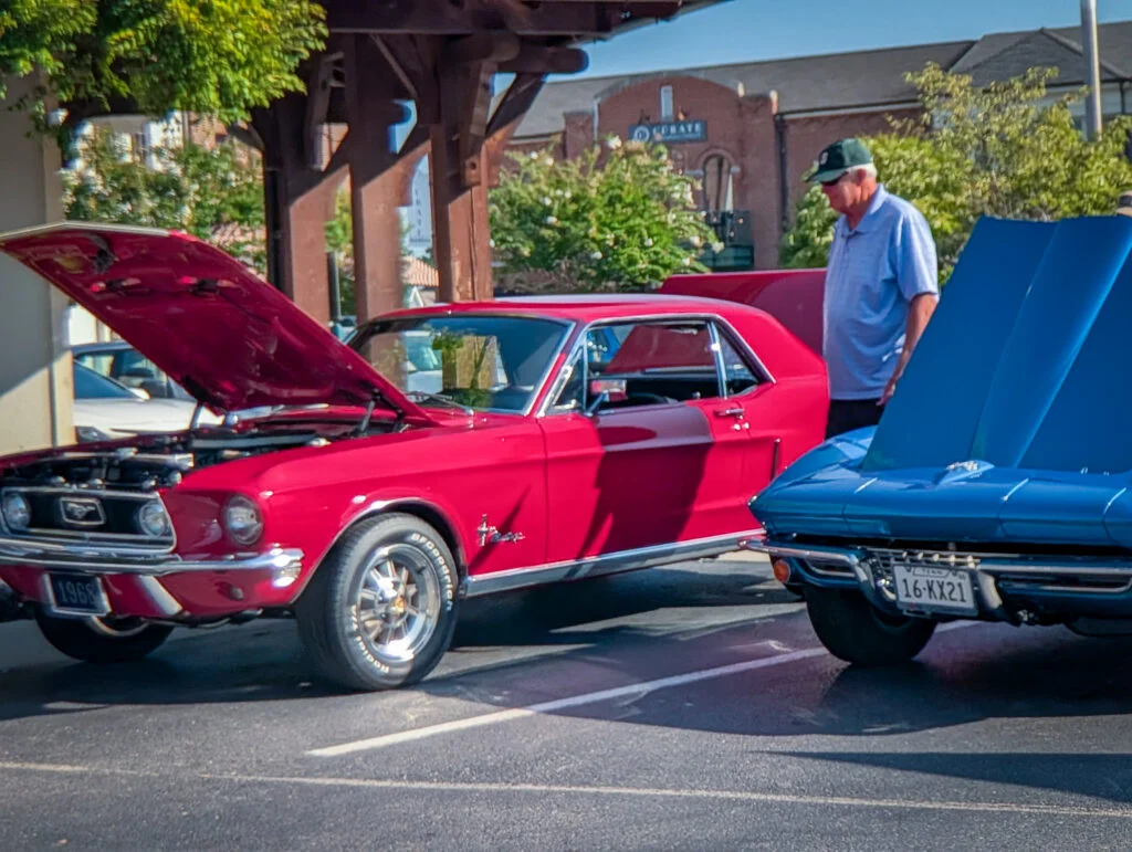 Red 1966 Ford Mustang with open hood and man inspecting it next to a blue car at an outdoor car show.