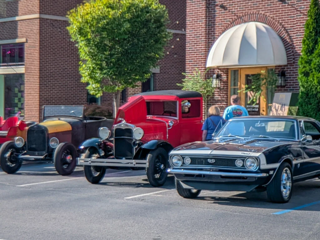 Row of vintage and classic cars parked outside a brick building with people nearby at a car event