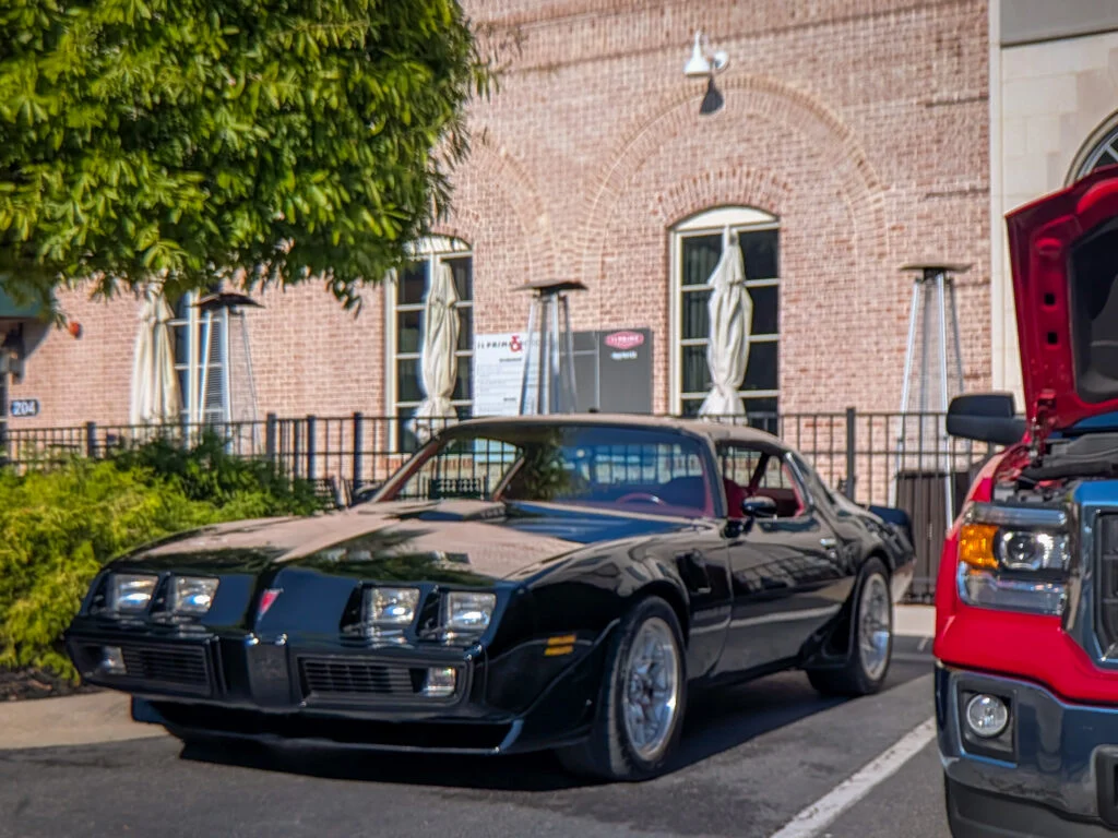 Black Pontiac Firebird parked on street with brick building and greenery in background during daytime