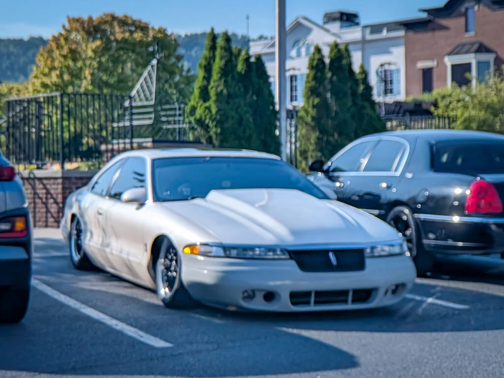 White Lincoln Mark VIII parked in a lot with trees and buildings in the background under clear sky