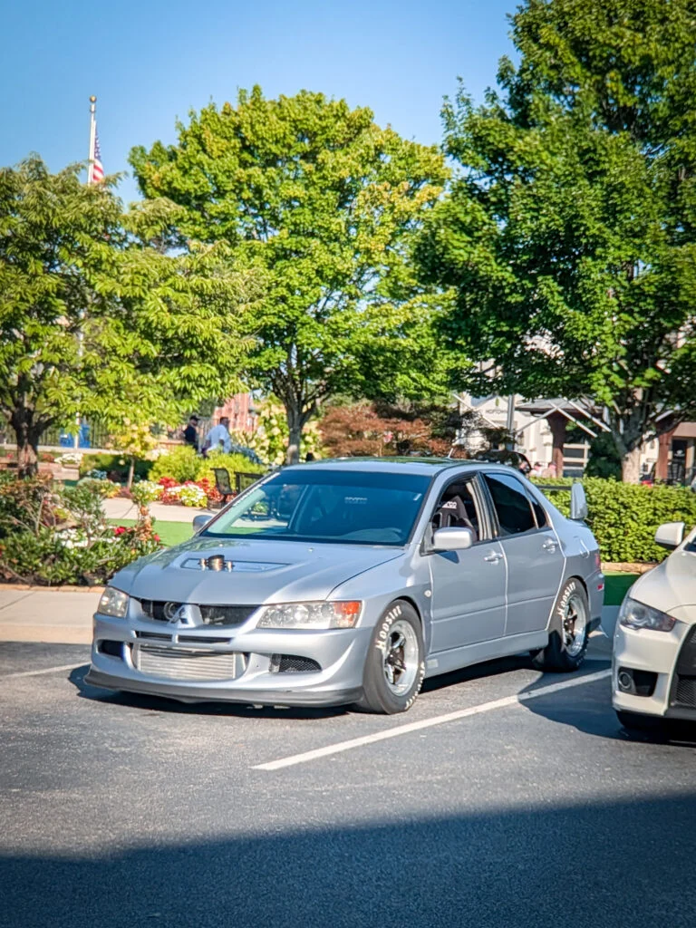 Silver Mitsubishi Lancer Evolution with aftermarket wheels and hood vents parked in a lot under trees on a sunny day