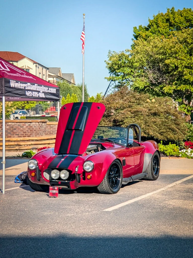 Red Shelby Cobra with black racing stripes and open hood in parking lot near a roofing company tent and American flag
