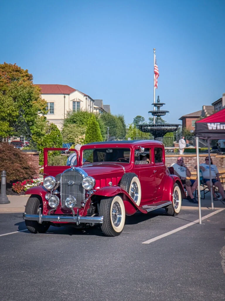 Red Buick Series 90 with whitewall tires parked in front of a fountain at a car event