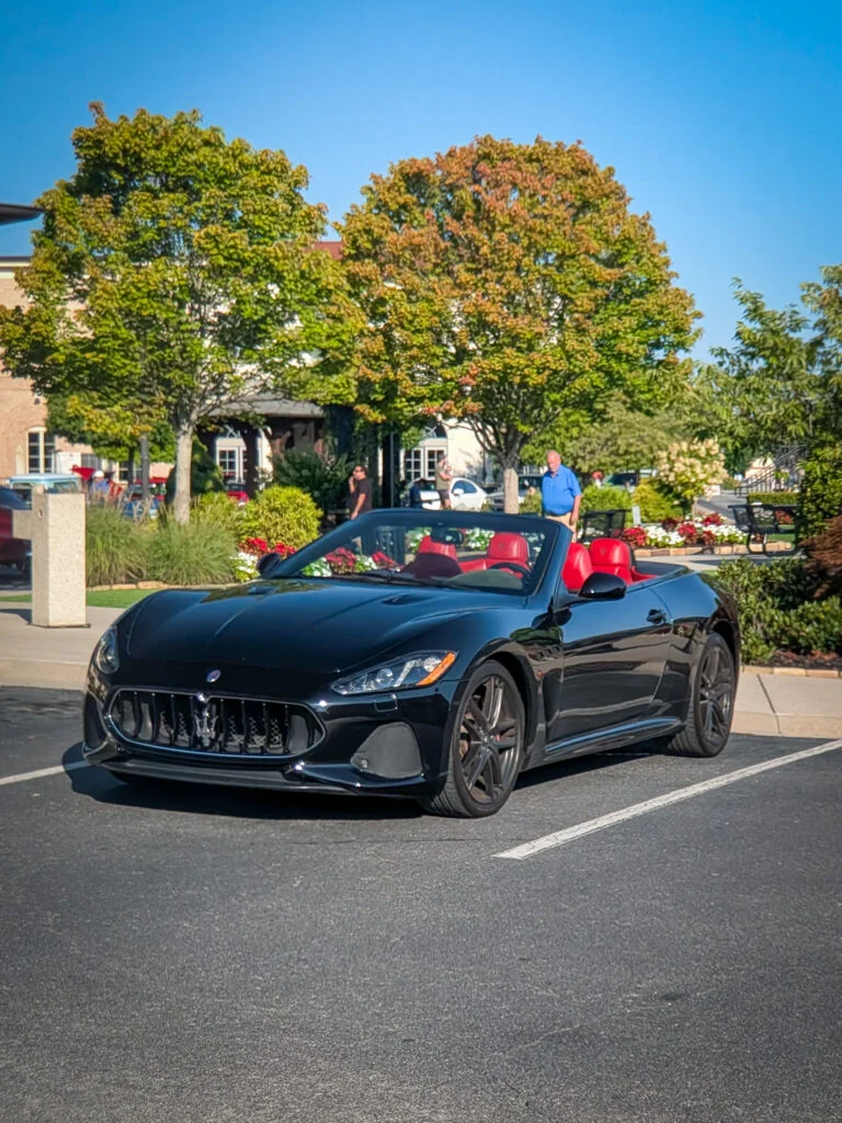 Black Maserati convertible with red interior parked near trees and people in an outdoor setting