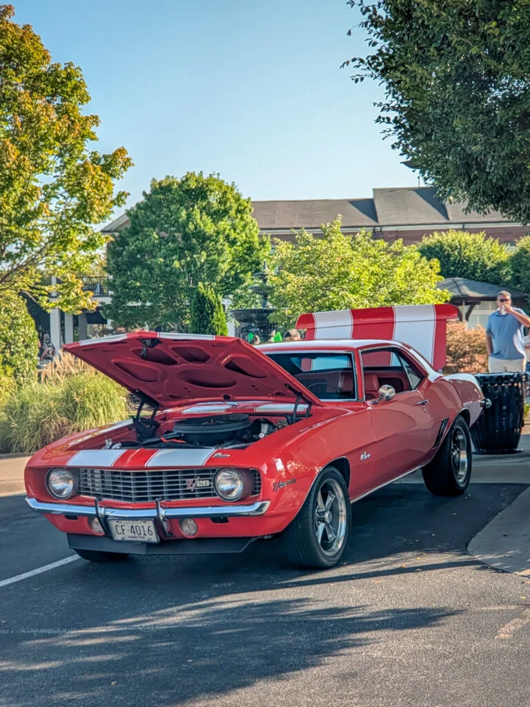 Red Chevrolet Camaro Z/28 with white racing stripes parked outdoors with hood open and trees in background