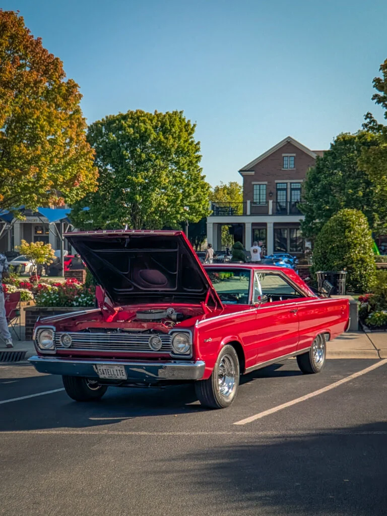 Red Dodge Dart with raised hood parked outdoors at a car event with greenery and a brick building in the background.