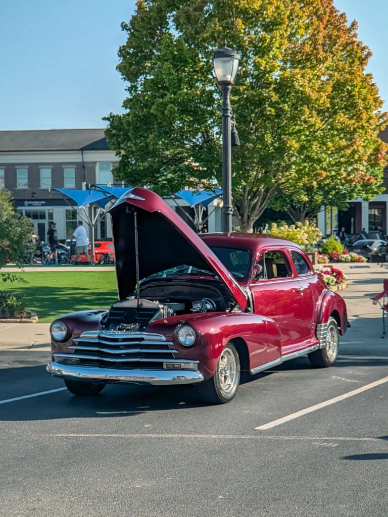 Maroon Chevrolet Stylemaster with hood open parked outdoors near greenery and buildings on a sunny day