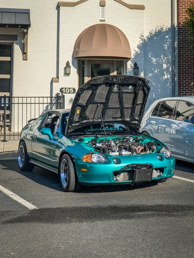 Teal-green Honda Del Sol with open hood showing engine, parked in front of building at car event