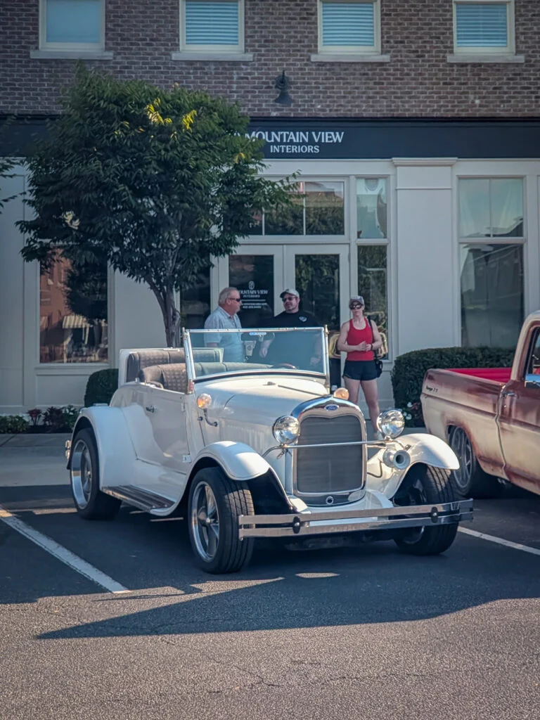 White vintage Ford Model A Roadster parked with three people standing nearby in front of a building with 'Mountain View Interiors'
