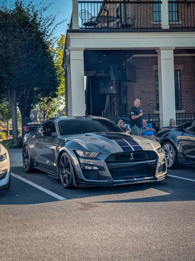 Silver Ford Mustang Shelby GT500 with black racing stripes parked in front of a brick building with people in background
