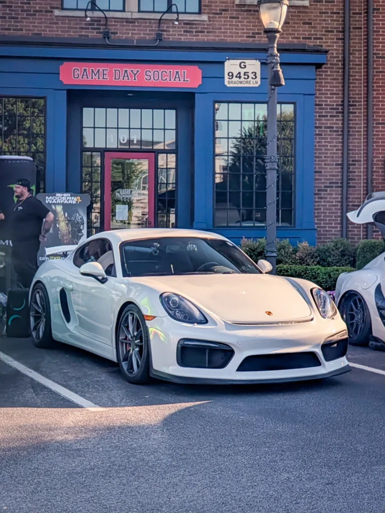 White Porsche Cayman GT4 parked in front of a brick building with blue trim and a Game Day Social sign