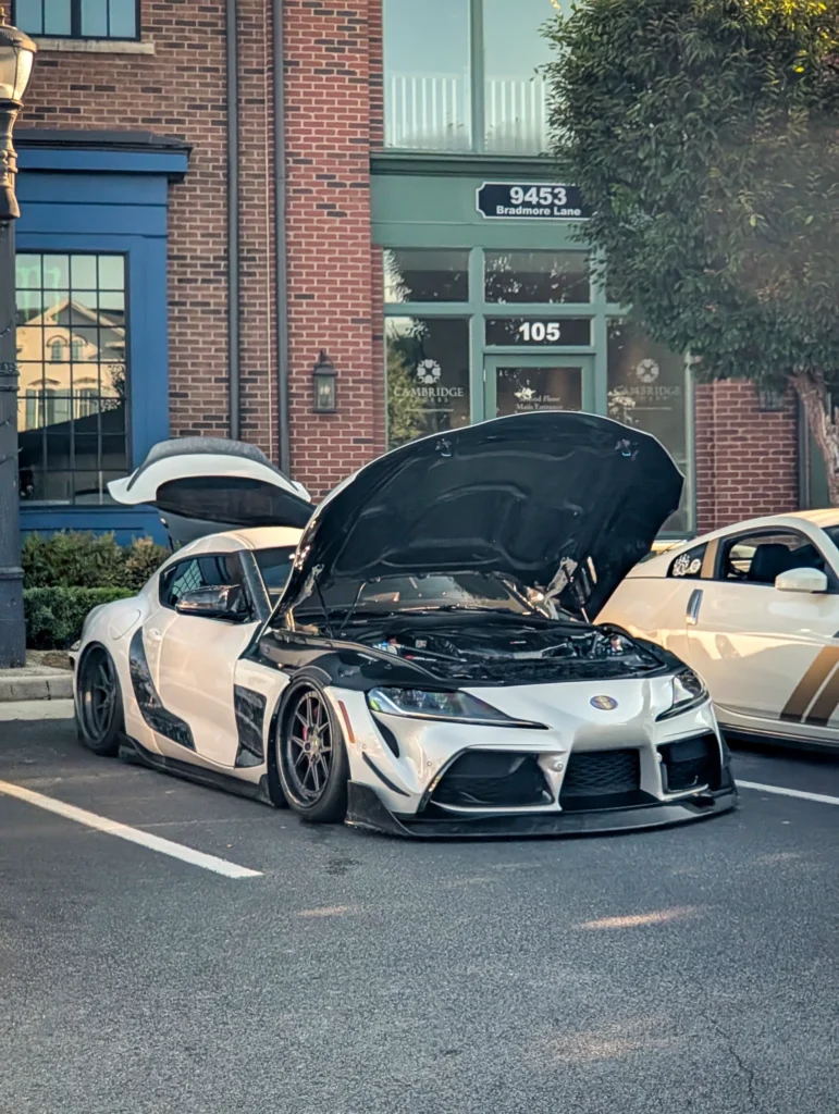 White Toyota Supra with black hood and fenders, hood and trunk open, parked in front of brick building