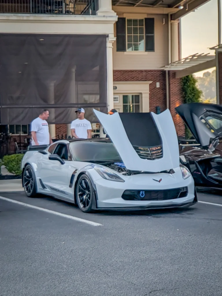 White Chevrolet Corvette with hood open parked in front of a building with two men standing nearby
