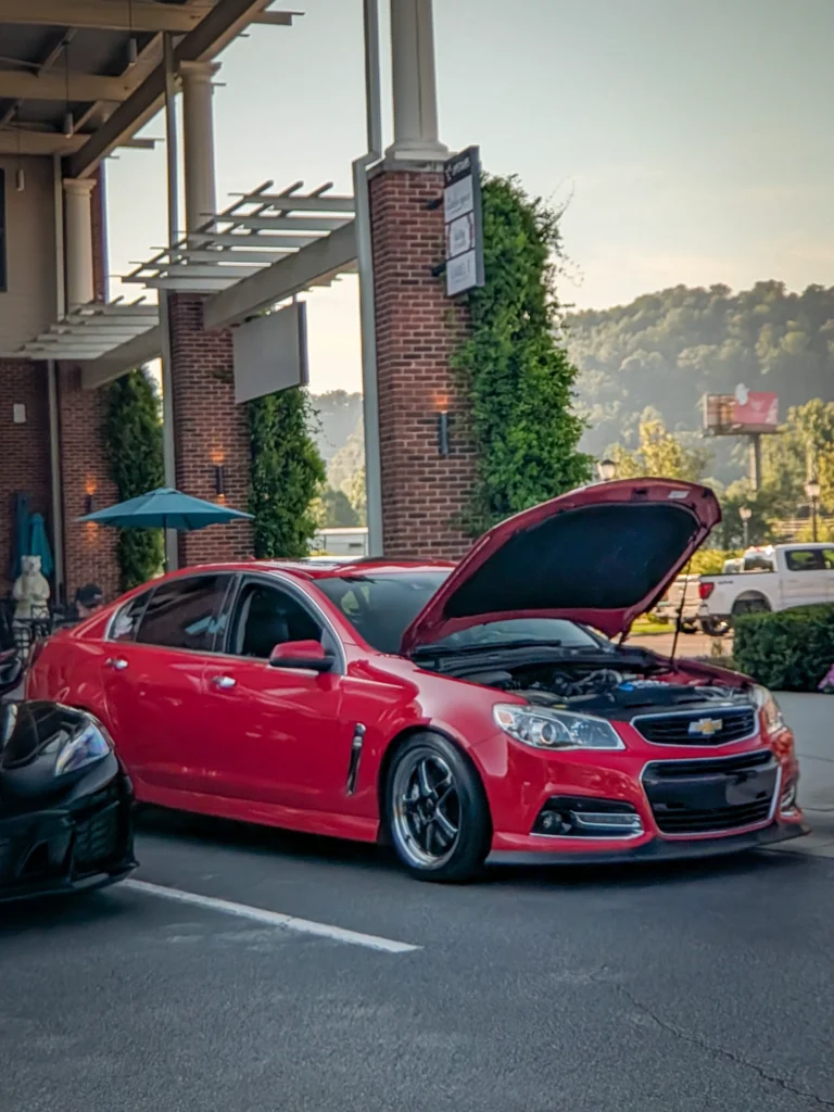 Red Chevrolet SS with hood open parked outside a brick building with greenery and mountains in background
