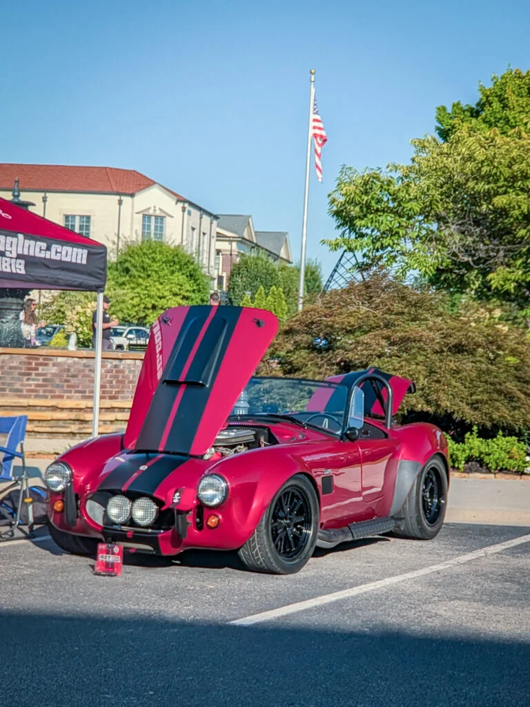 Red Shelby Cobra with black racing stripes, hood and trunk open, parked at an outdoor car event under clear sky